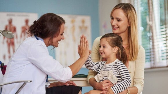 Uma médica com estetoscópio sorri e faz um "high-five" com uma menina pequena, que sorri de volta enquanto está sentada no colo de sua mãe, que também sorri. Uma médica com estetoscópio sorri e faz um "high-five" com uma menina pequena, que sorri de volta enquanto está sentada no colo de sua mãe, que também sorri.