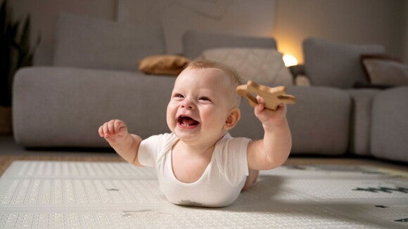 Um bebê deitado de barriga para baixo em um tapete educativo, segurando um brinquedo de madeira e sorrindo. Um bebê deitado de barriga para baixo em um tapete educativo, segurando um brinquedo de madeira e sorrindo.