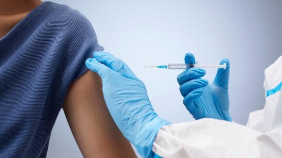 Close-up de um profissional de saúde, com luvas e jaleco de proteção, aplicando uma injeção no braço de uma pessoa.