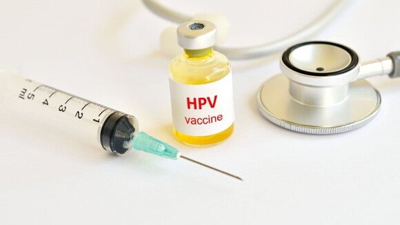 Frasco de vidro contendo a vacina HPV (com a etiqueta "HPV vaccine"), uma seringa e um estetoscópio sobre uma superfície branca, representando a prevenção.