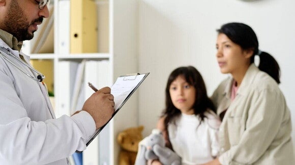 Uma mãe e sua filha em uma consulta médica, enquanto o médico anota informações em um prontuário. A imagem ilustra o diagnóstico e tratamento de verme infantil.