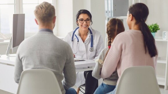 Uma médica pediatra, com um jaleco branco e um estetoscópio, sorri enquanto conversa com um casal e sua filha, que aparecem de costas na imagem. A família está sentada à frente da mesa da médica. 