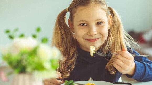 Uma menina sentada à uma mesa, com um prato de comida à sua frente, sorrindo enquanto se alimenta. Uma menina sentada à uma mesa, com um prato de comida à sua frente, sorrindo enquanto se alimenta.