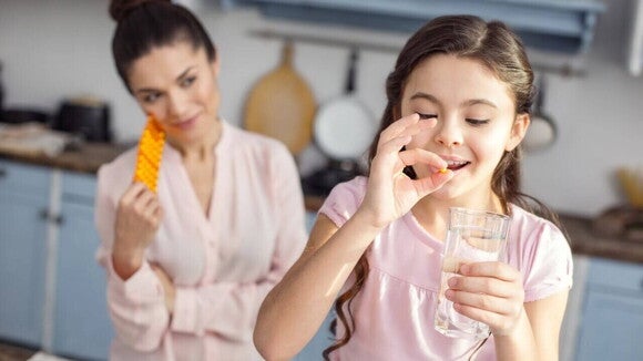 Vitaminas para criança Uma menina em uma cozinha iluminada, tomando goma de vitamina com um copo de água. Uma mulher adulta a observa por cima do ombro, segurando um blister de vitaminas.
