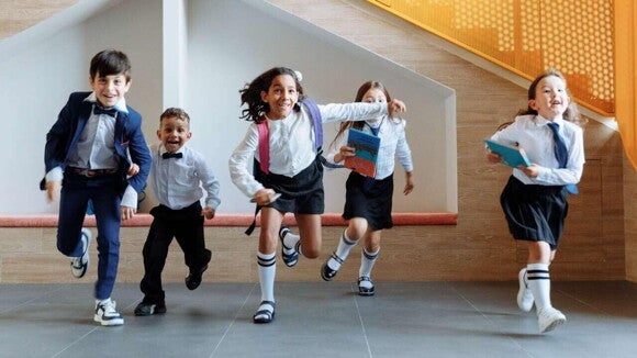 Um grupo de cinco crianças, vestindo uniformes escolares, corre alegremente pelo corredor de uma escola. A expressão de entusiasmo e felicidade em seus rostos sugere a empolgação com o início do ano letivo. A imagem captura a alegria e a energia que a volta às aulas traz para os alunos.